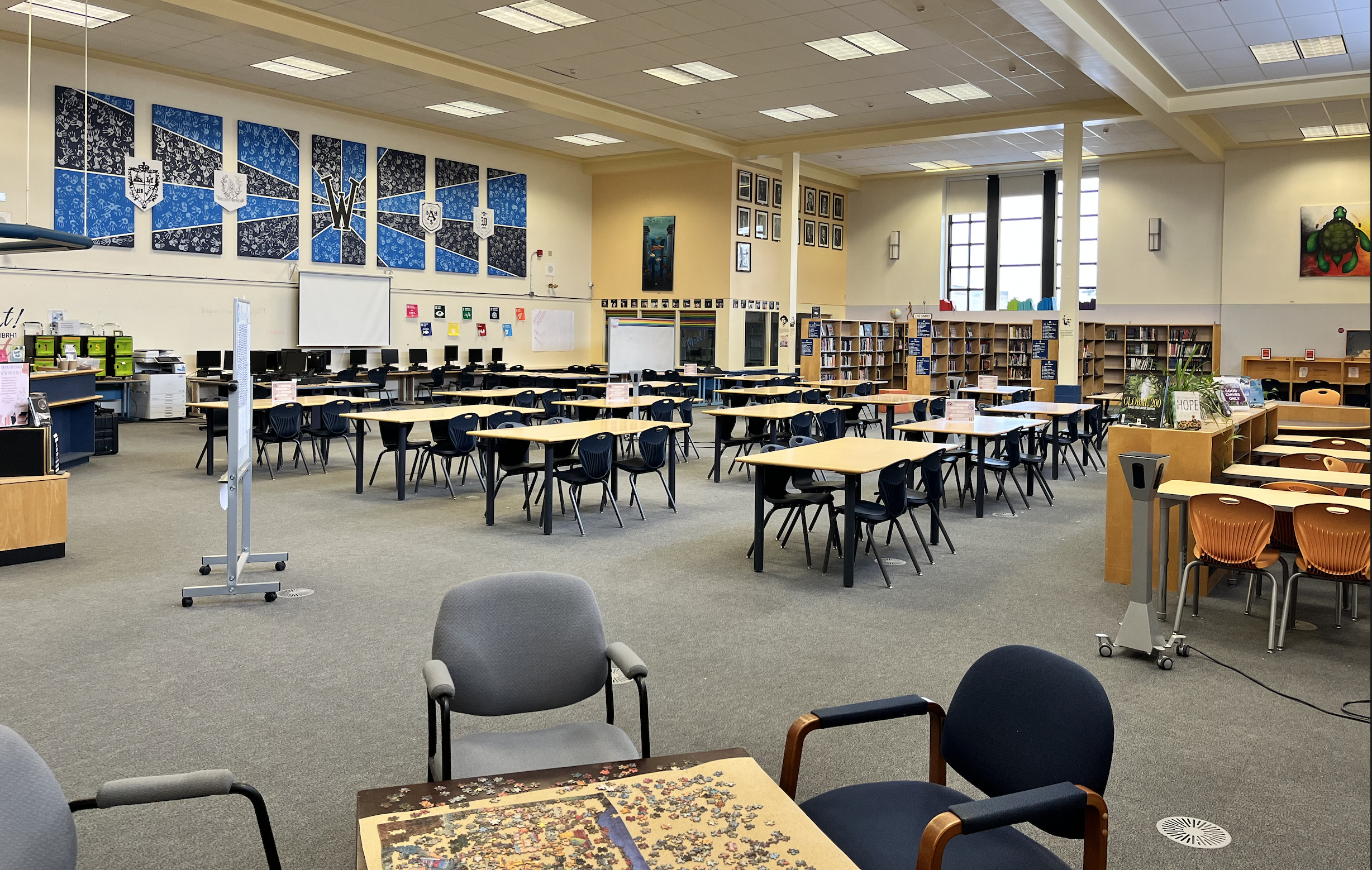 Room full of tables and chairs, a jigsaw puzzle on a table close to the camera. A library desk is in the corner.